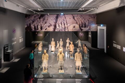 Wide installation view of the Terracotta Warriors displayed in a glass enclosure beneath a panoramic image of the excavation pits.