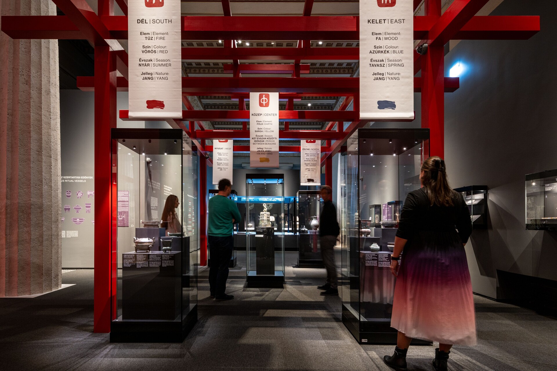 Gallery view with a red architectural framework, suspended bilingual five-elements banners, visitors, and archaeological objects in glass cases.