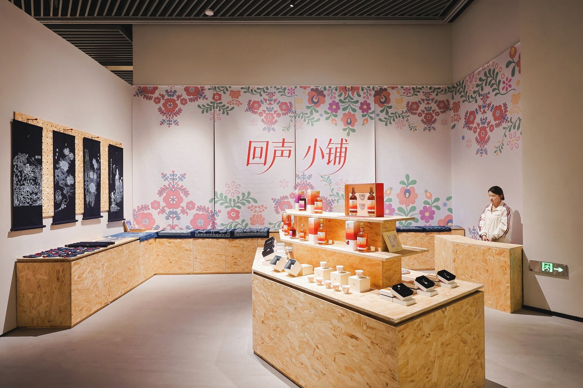 Installation view of “Songs of Life Along the Danube: An Exhibition of Hungarian Intangible Cultural Heritage” in Hangzhou, showing plywood display platforms, floral folk-pattern wall graphics, black textile panels, bottles and small display objects, with a seated visitor or staff member at right.