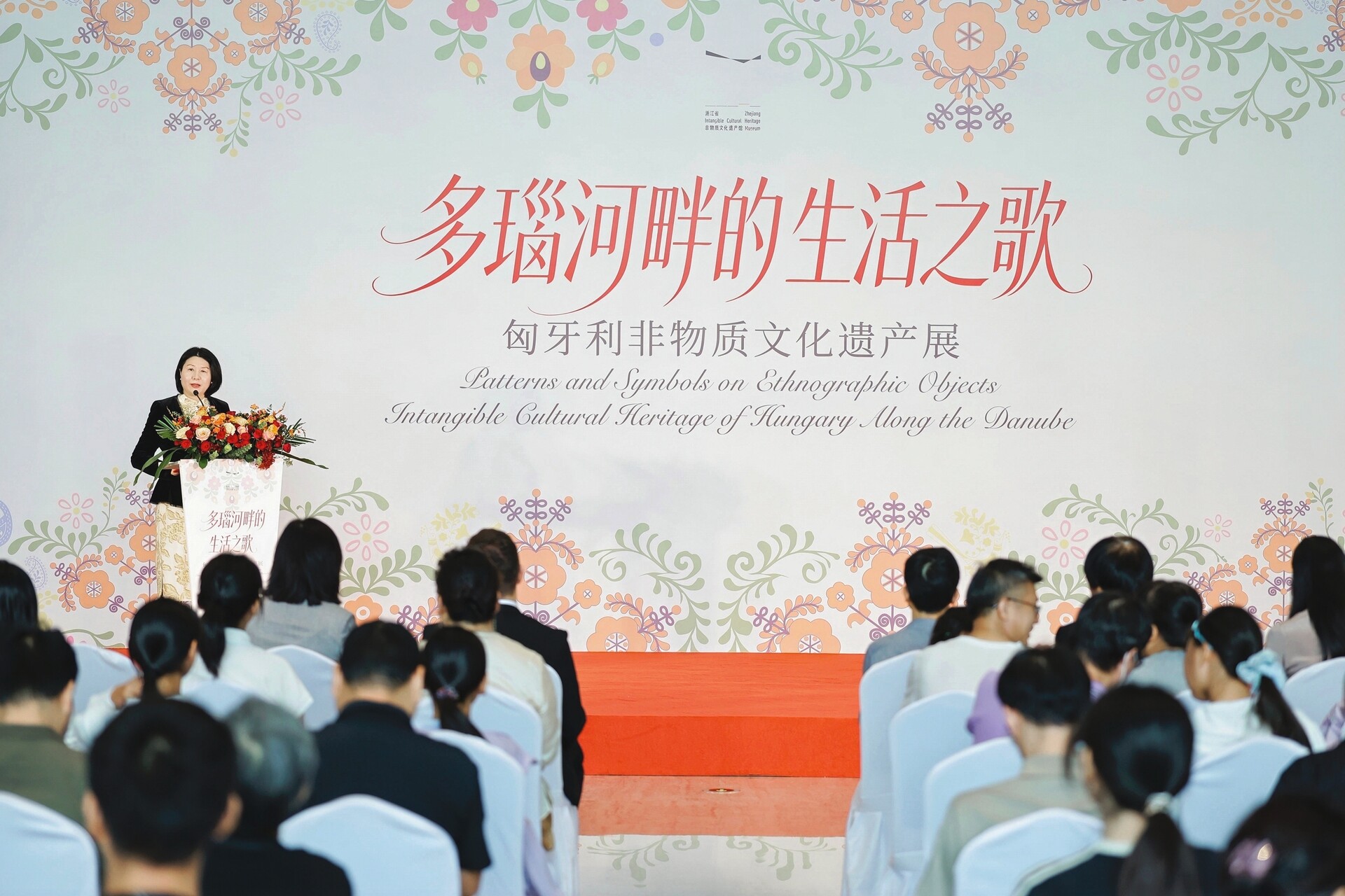 Director Guo Yi speaks at a flower-adorned lectern before a seated audience, with a floral backdrop for the exhibition “Songs of Life Along the Danube: An Exhibition of Hungarian Intangible Cultural Heritage” at the Zhejiang Intangible Cultural Heritage Museum.