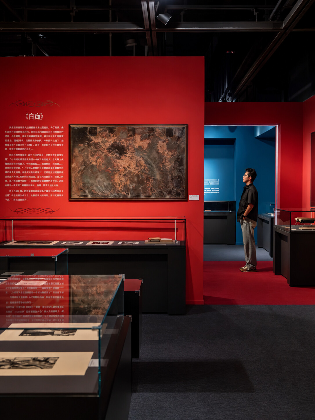 “陀思妥耶夫斯基的世界：停止阅读意味着停止思考”展览现场，红色展区展示《白痴》相关内容，墙上悬挂一件装框作品，展柜中陈列印刷品，一名观众站在通向蓝色展厅的入口旁。